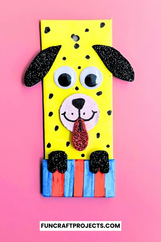 Cardboard Dog Bookmark Craft is a dog-themed bookmark made from yellow cardboard, featuring black glitter ears, googly eyes, and a red tongue, with a striped blue and red collar.