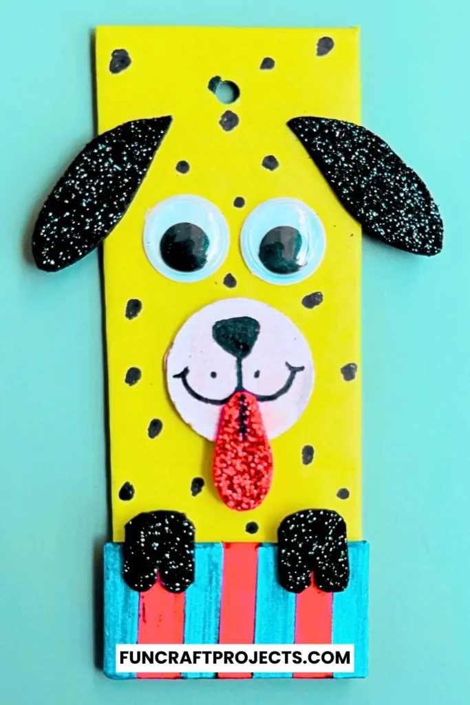 Cardboard Dog Bookmark Craft is a dog-themed bookmark made from yellow cardboard, featuring black glitter ears, googly eyes, and a red tongue, with a striped blue and red collar.