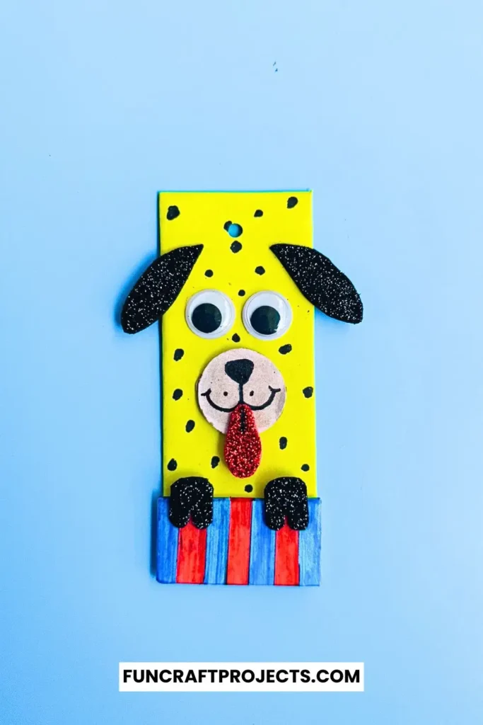 Cardboard Dog Bookmark Craft is a dog-themed bookmark made from yellow cardboard, featuring black glitter ears, googly eyes, and a red tongue, with a striped blue and red collar.