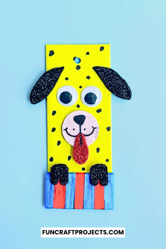 Cardboard Dog Bookmark Craft is a dog-themed bookmark made from yellow cardboard, featuring black glitter ears, googly eyes, and a red tongue, with a striped blue and red collar.