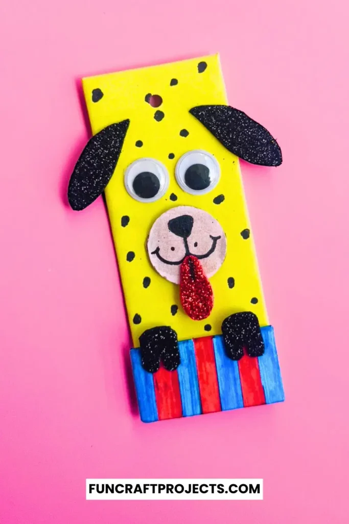 Cardboard Dog Bookmark Craft is a dog-themed bookmark made from yellow cardboard, featuring black glitter ears, googly eyes, and a red tongue, with a striped blue and red collar.