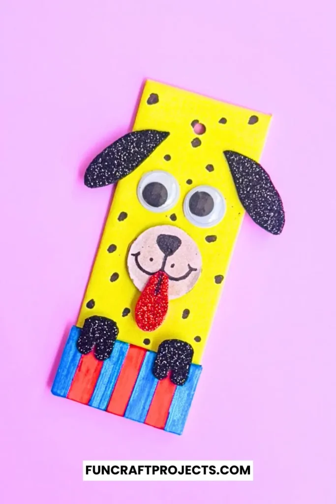 Cardboard Dog Bookmark Craft is a dog-themed bookmark made from yellow cardboard, featuring black glitter ears, googly eyes, and a red tongue, with a striped blue and red collar.