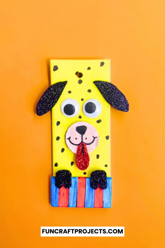 Cardboard Dog Bookmark Craft is a dog-themed bookmark made from yellow cardboard, featuring black glitter ears, googly eyes, and a red tongue, with a striped blue and red collar.