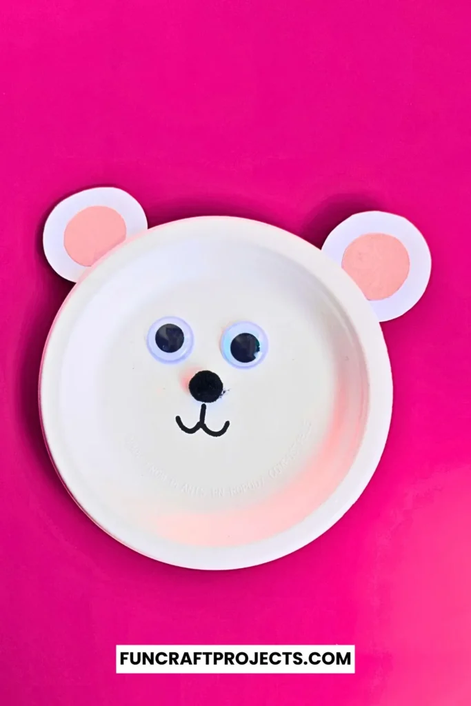 Paper plate polar bear craft with googly eyes, pink ears, and a simple black nose. Easy DIY bear craft for kids, perfect for preschool or winter activities.