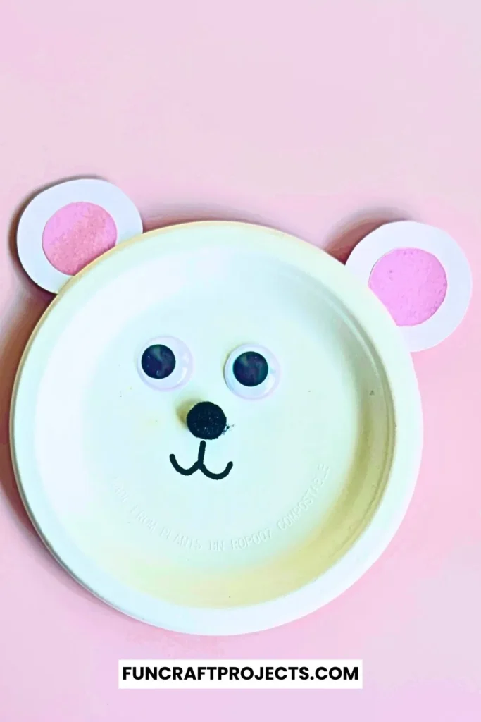 Paper plate polar bear craft with googly eyes, pink ears, and a simple black nose. Easy DIY bear craft for kids, perfect for preschool or winter activities.