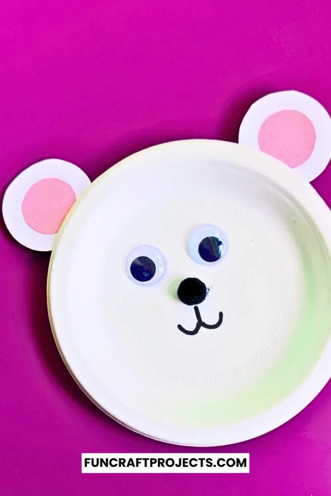 Paper plate polar bear craft with googly eyes, pink ears, and a simple black nose. Easy DIY bear craft for kids, perfect for preschool or winter activities.