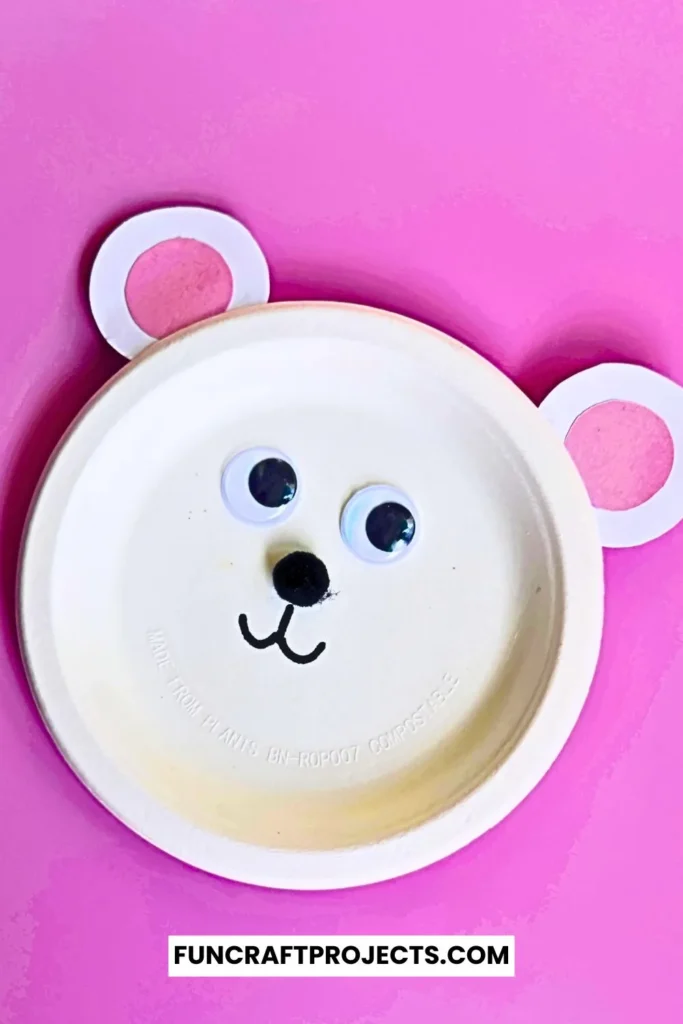 Paper plate polar bear craft with googly eyes, pink ears, and a simple black nose. Easy DIY bear craft for kids, perfect for preschool or winter activities.