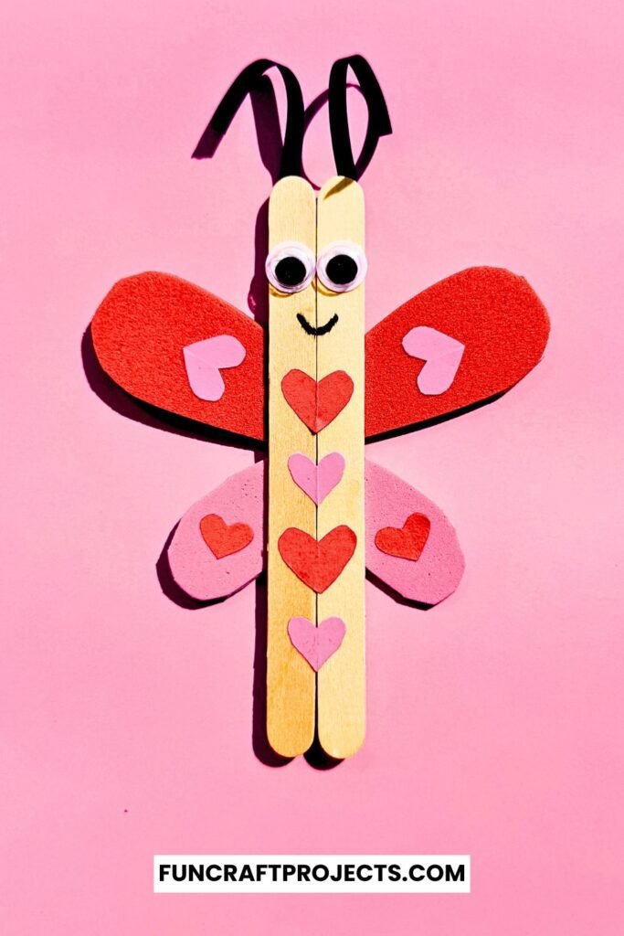 A cute Popsicle Stick Love Bug Craft made with wooden craft sticks, red and pink heart-shaped wings, and googly eyes, displayed on a blue background.