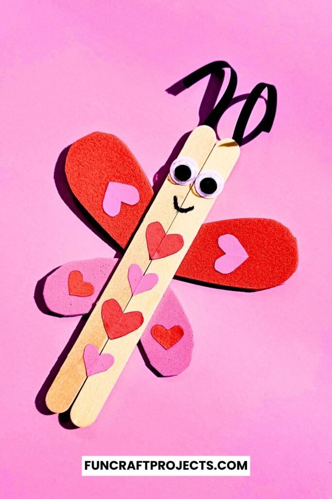 A cute Popsicle Stick Love Bug Craft made with wooden craft sticks, red and pink heart-shaped wings, and googly eyes, displayed on a blue background.