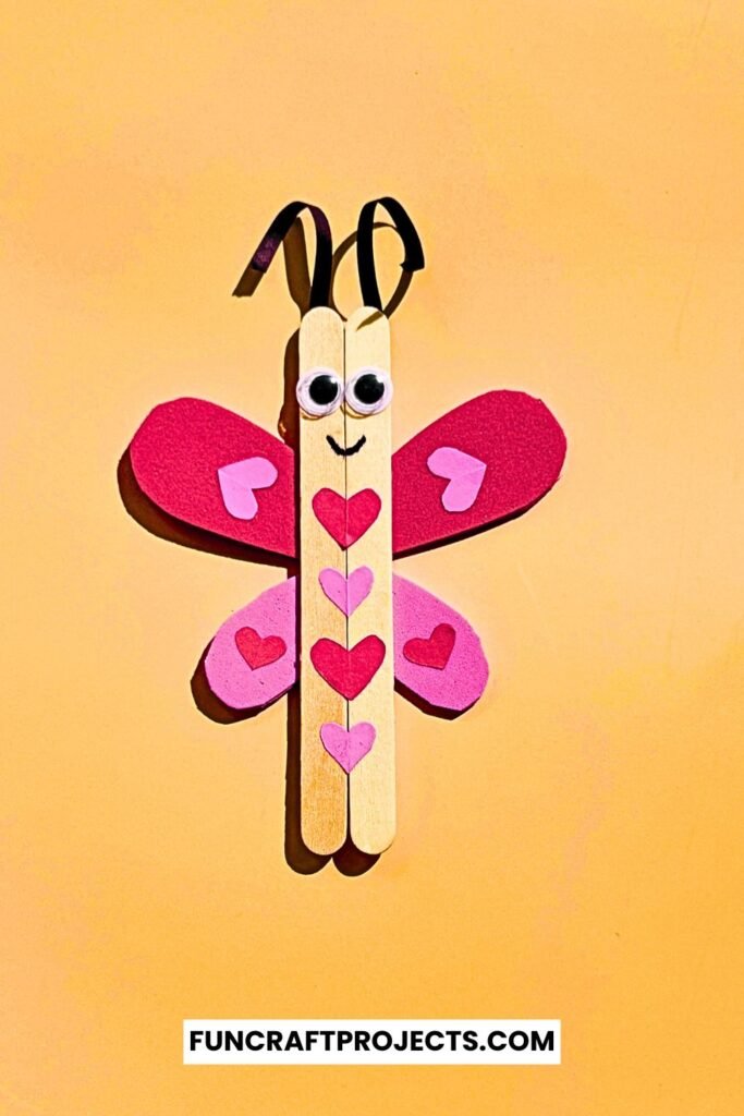 A cute Popsicle Stick Love Bug Craft made with wooden craft sticks, red and pink heart-shaped wings, and googly eyes, displayed on a blue background.