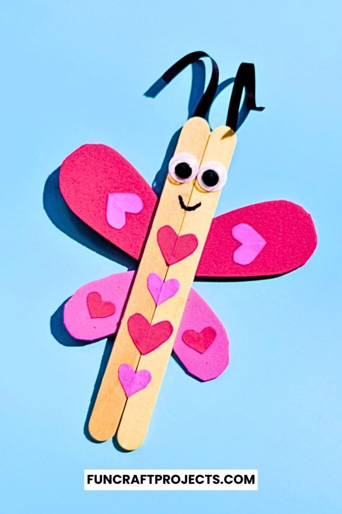 A cute Popsicle Stick Love Bug Craft made with wooden craft sticks, red and pink heart-shaped wings, and googly eyes, displayed on a blue background.