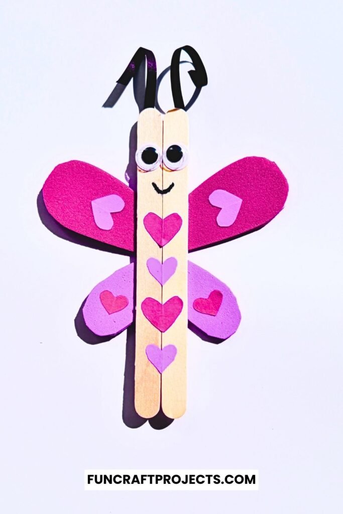 A cute Popsicle Stick Love Bug Craft made with wooden craft sticks, red and pink heart-shaped wings, and googly eyes, displayed on a blue background.