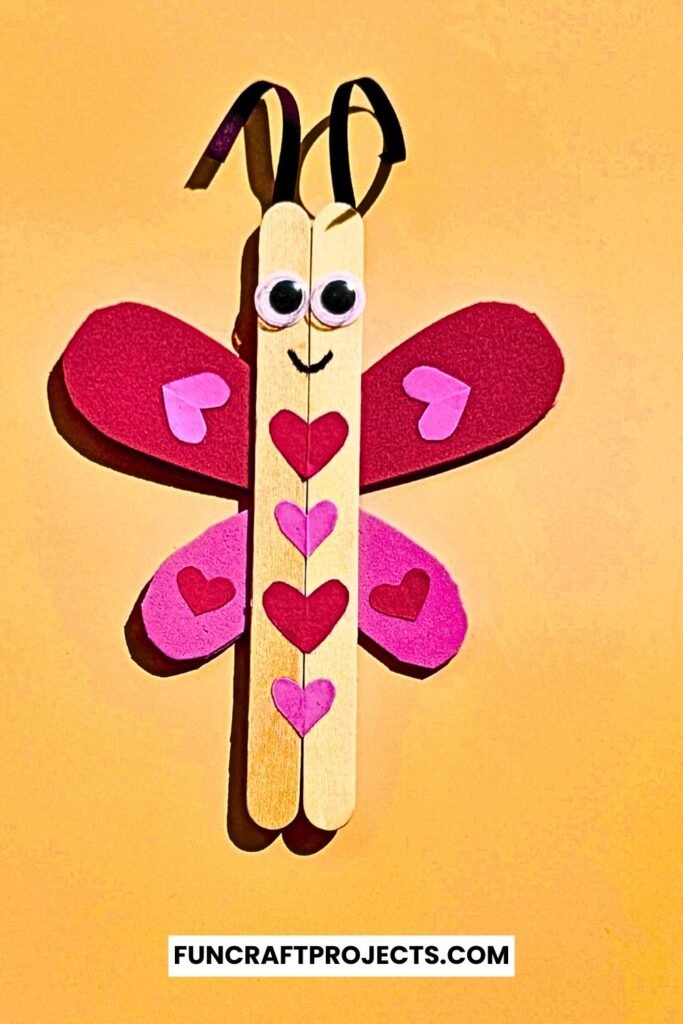 A cute Popsicle Stick Love Bug Craft made with wooden craft sticks, red and pink heart-shaped wings, and googly eyes, displayed on a blue background.