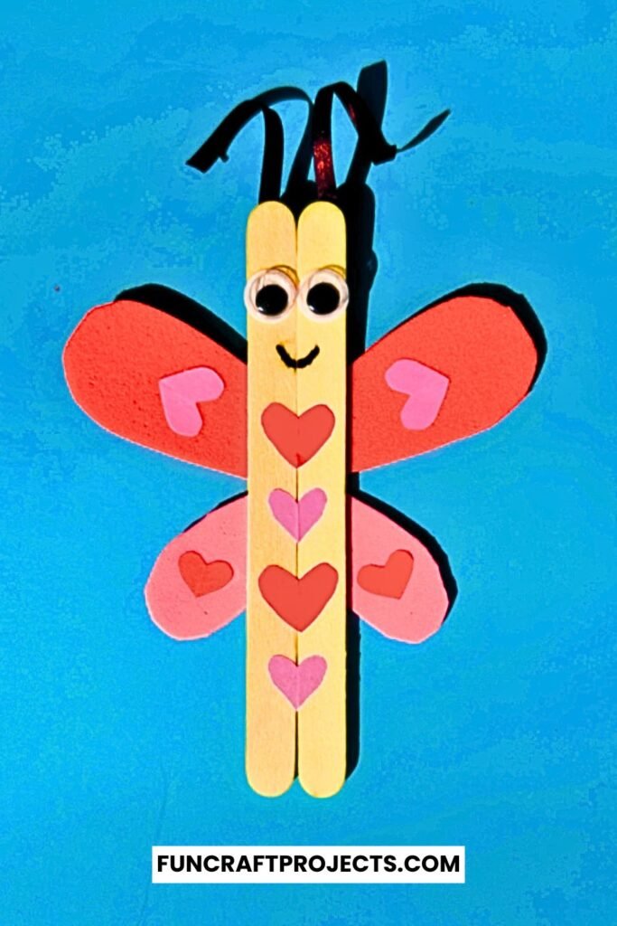 A cute Popsicle Stick Love Bug Craft made with wooden craft sticks, red and pink heart-shaped wings, and googly eyes, displayed on a blue background.