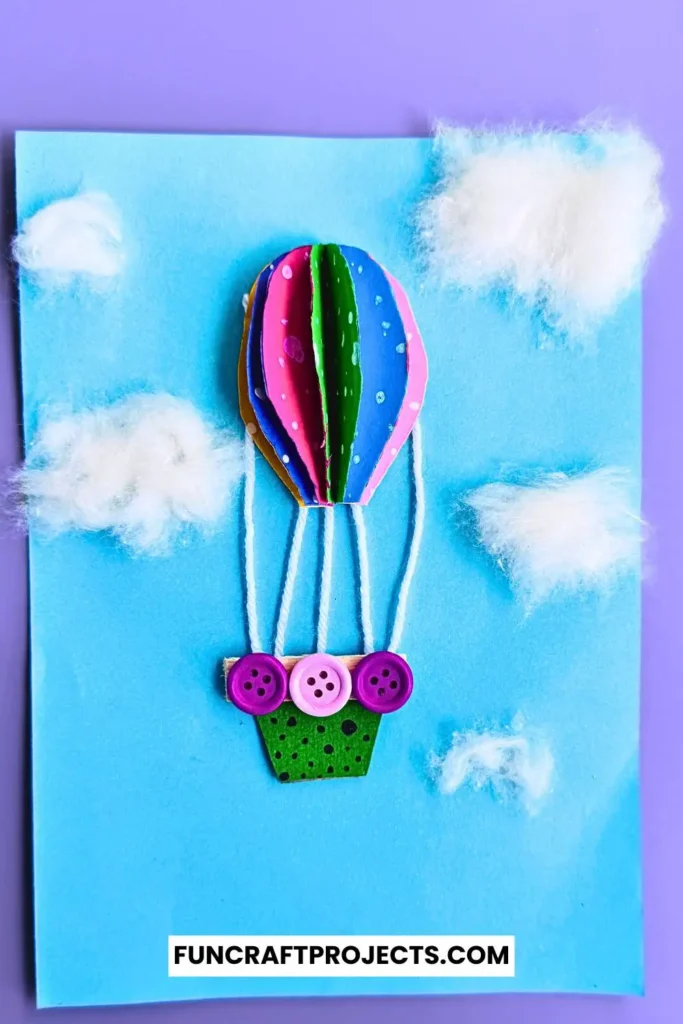 Bright and colorful construction paper hot air balloon craft with 3D effects, buttons for a basket, and fluffy cotton clouds on a blue background