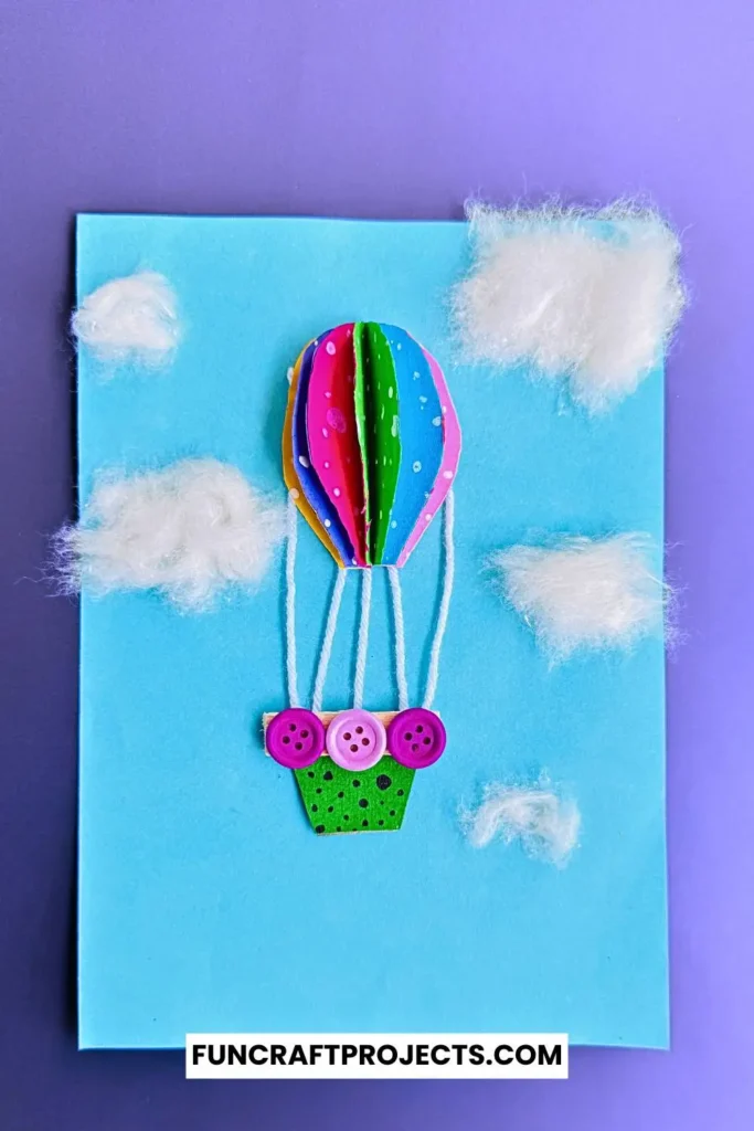 Bright and colorful construction paper hot air balloon craft with 3D effects, buttons for a basket, and fluffy cotton clouds on a blue background