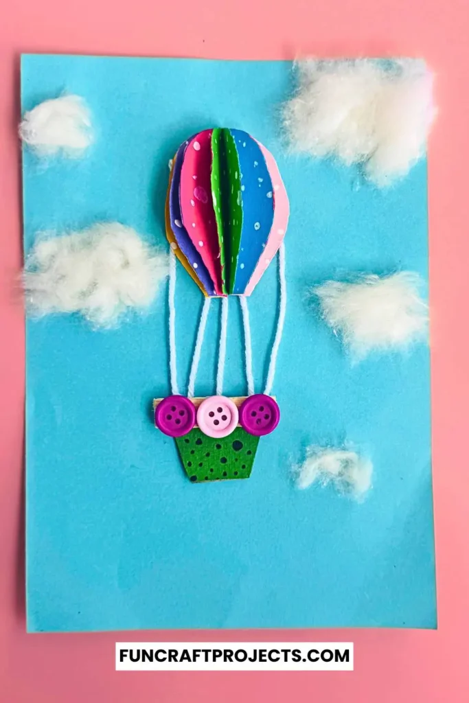 Bright and colorful construction paper hot air balloon craft with 3D effects, buttons for a basket, and fluffy cotton clouds on a blue background