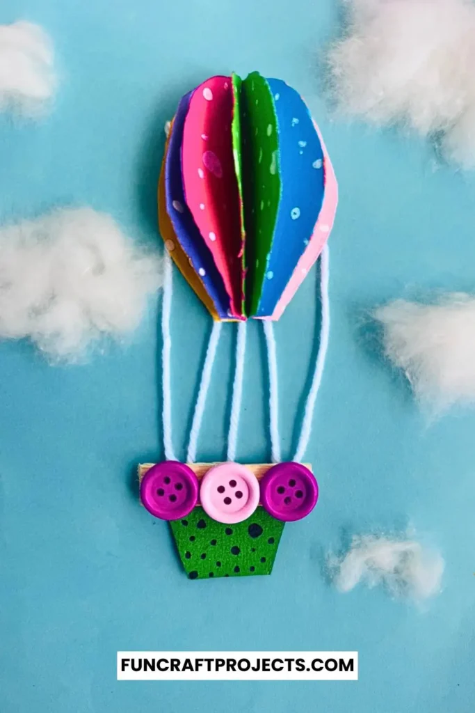 Bright and colorful construction paper hot air balloon craft with 3D effects, buttons for a basket, and fluffy cotton clouds on a blue background