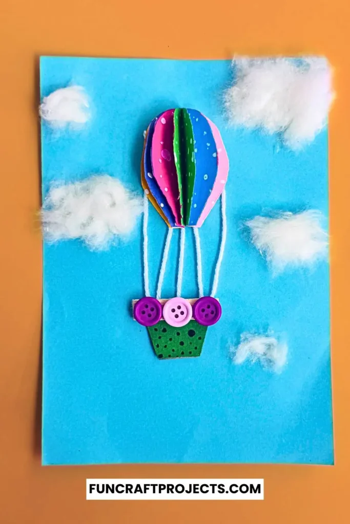 Bright and colorful construction paper hot air balloon craft with 3D effects, buttons for a basket, and fluffy cotton clouds on a blue background