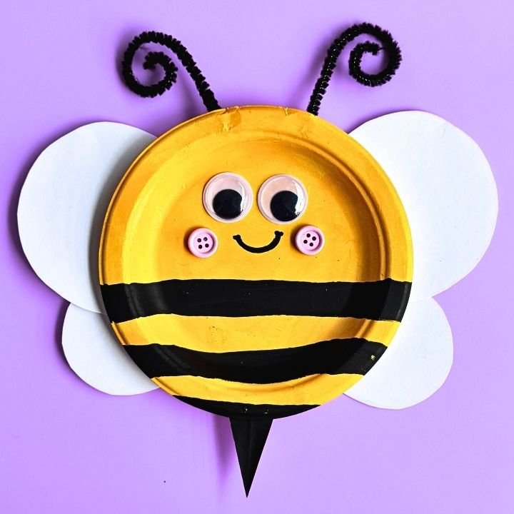 Bright and cheerful paper plate honey bee craft with yellow and black stripes, googly eyes, pipe cleaner antennae, and white paper wings on a blue background.