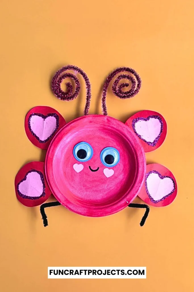 Paper plate love bug craft with pink paint, heart-shaped wings, googly eyes, and pipe cleaner antennae on a beige background.