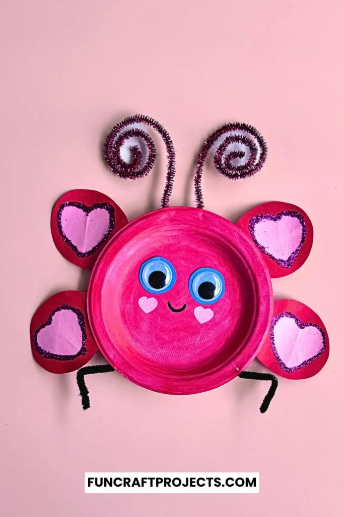 Paper plate love bug craft with pink paint, heart-shaped wings, googly eyes, and pipe cleaner antennae on a beige background.