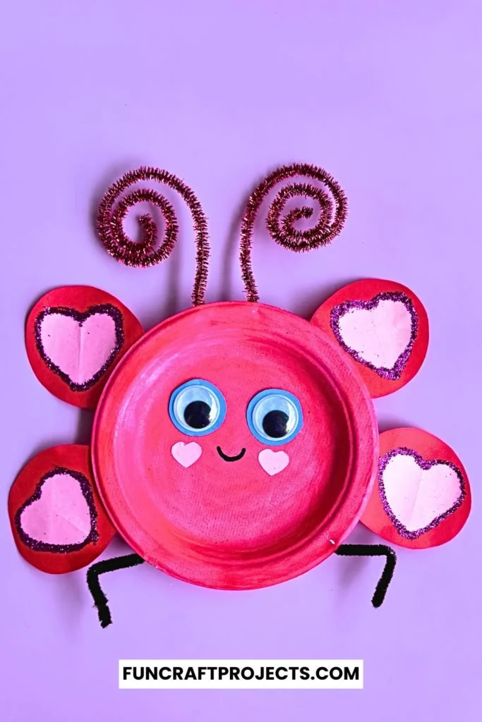 Paper plate love bug craft with pink paint, heart-shaped wings, googly eyes, and pipe cleaner antennae on a beige background.