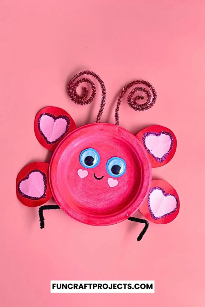 Paper plate love bug craft with pink paint, heart-shaped wings, googly eyes, and pipe cleaner antennae on a beige background.