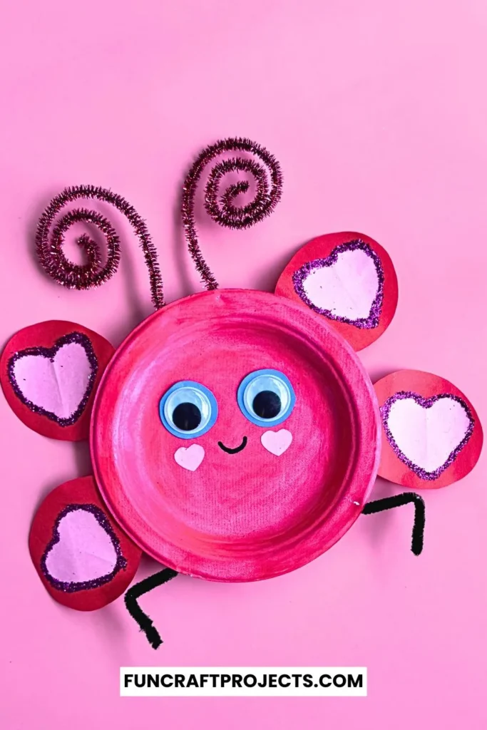 Paper plate love bug craft with pink paint, heart-shaped wings, googly eyes, and pipe cleaner antennae on a beige background.