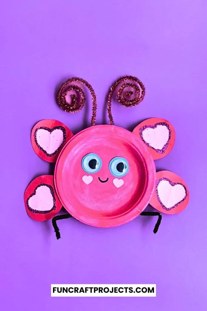 Paper plate love bug craft with pink paint, heart-shaped wings, googly eyes, and pipe cleaner antennae on a beige background.