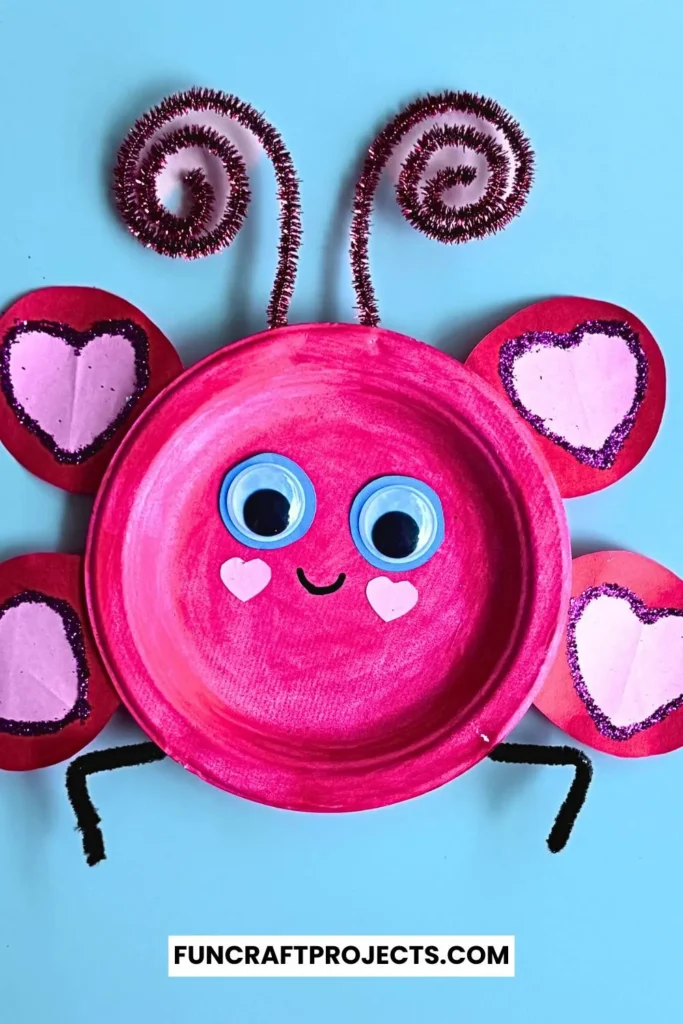 Paper plate love bug craft with pink paint, heart-shaped wings, googly eyes, and pipe cleaner antennae on a beige background.