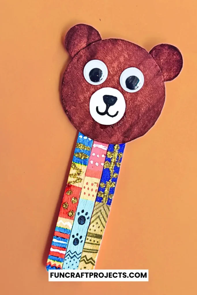 Handmade popsicle stick teddy bear bookmark craft with colorful patterns and googly eyes, crafted from jumbo popsicle sticks and brown craft paper