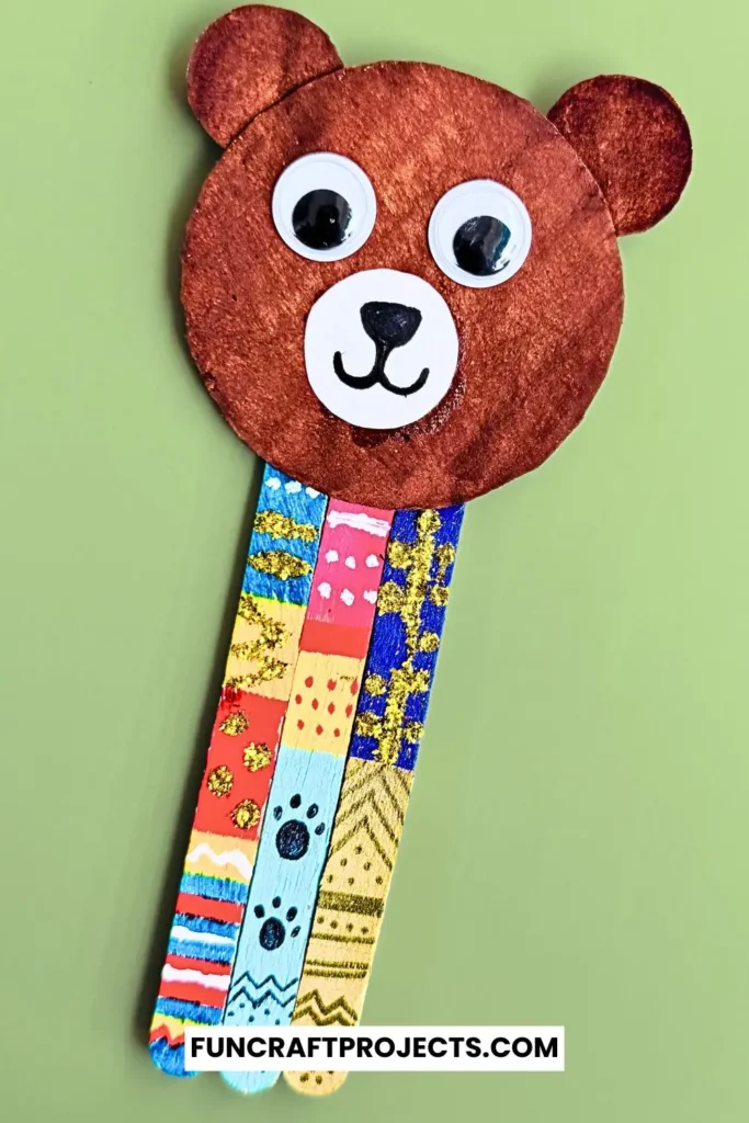 Handmade popsicle stick teddy bear bookmark craft with colorful patterns and googly eyes, crafted from jumbo popsicle sticks and brown craft paper