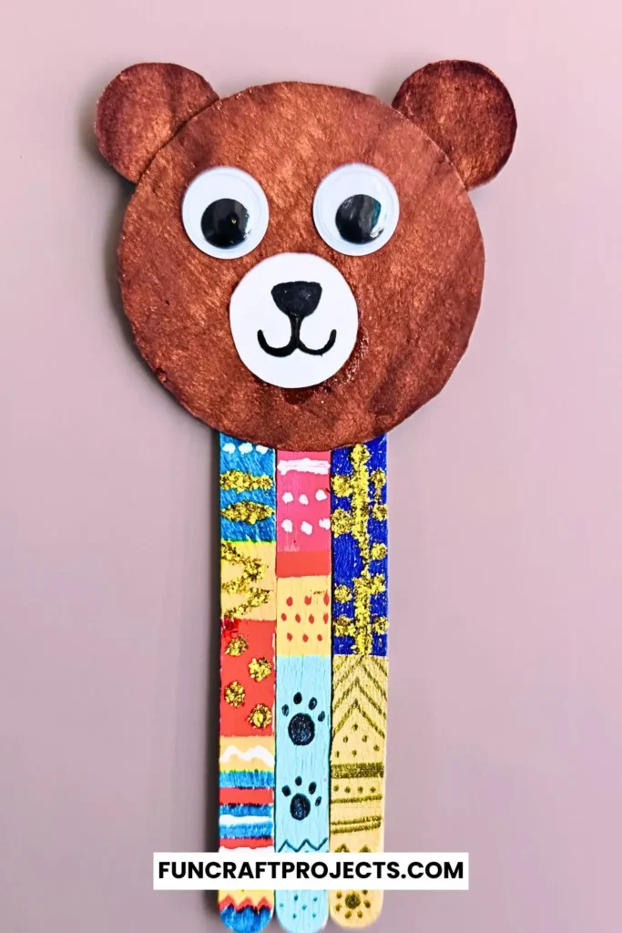 Handmade popsicle stick teddy bear bookmark craft with colorful patterns and googly eyes, crafted from jumbo popsicle sticks and brown craft paper