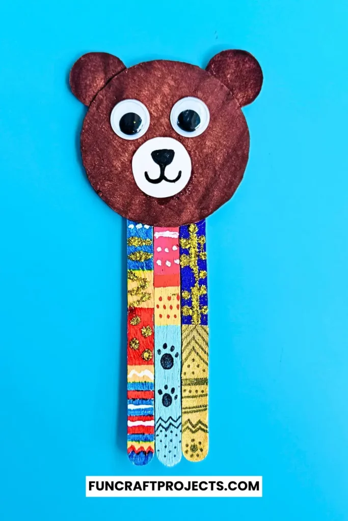 Handmade popsicle stick teddy bear bookmark craft with colorful patterns and googly eyes, crafted from jumbo popsicle sticks and brown craft paper