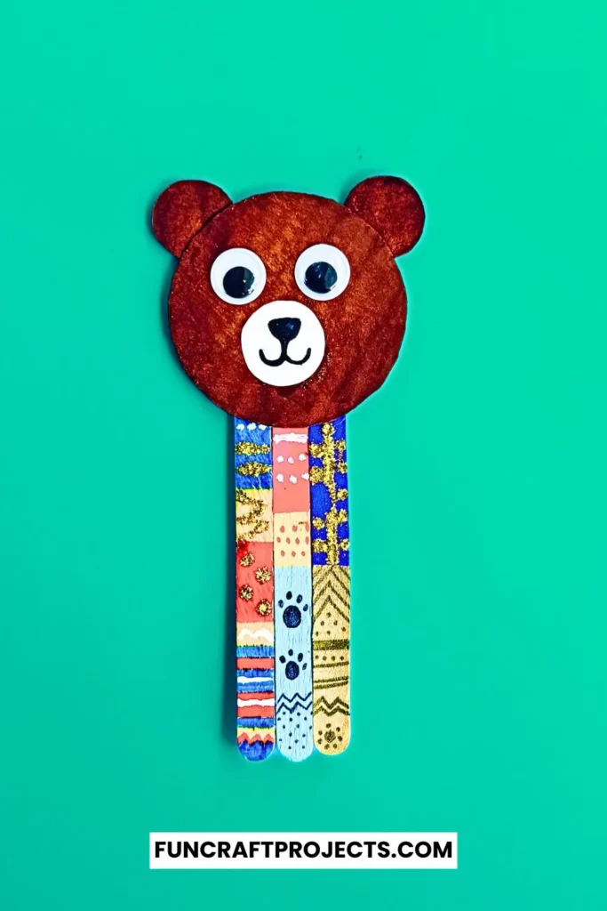 Handmade popsicle stick teddy bear bookmark craft with colorful patterns and googly eyes, crafted from jumbo popsicle sticks and brown craft paper