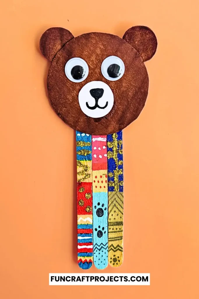 Handmade popsicle stick teddy bear bookmark craft with colorful patterns and googly eyes, crafted from jumbo popsicle sticks and brown craft paper