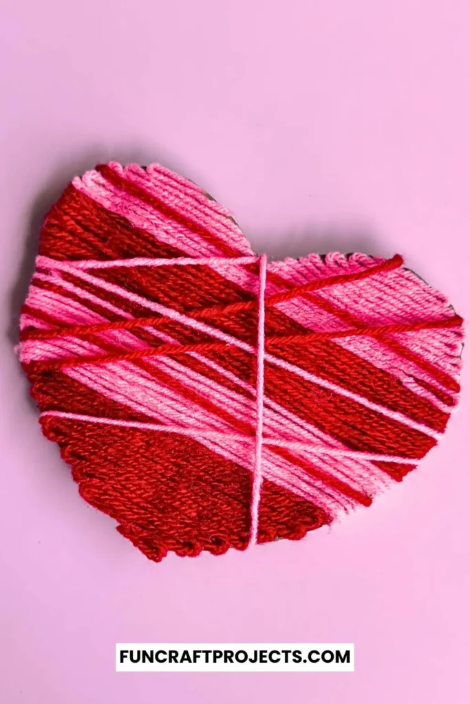 A vibrant yarn wrapped heart craft featuring pink and red yarn, wrapped intricately around a sturdy heart-shaped cardboard, perfect for Valentine’s Day.