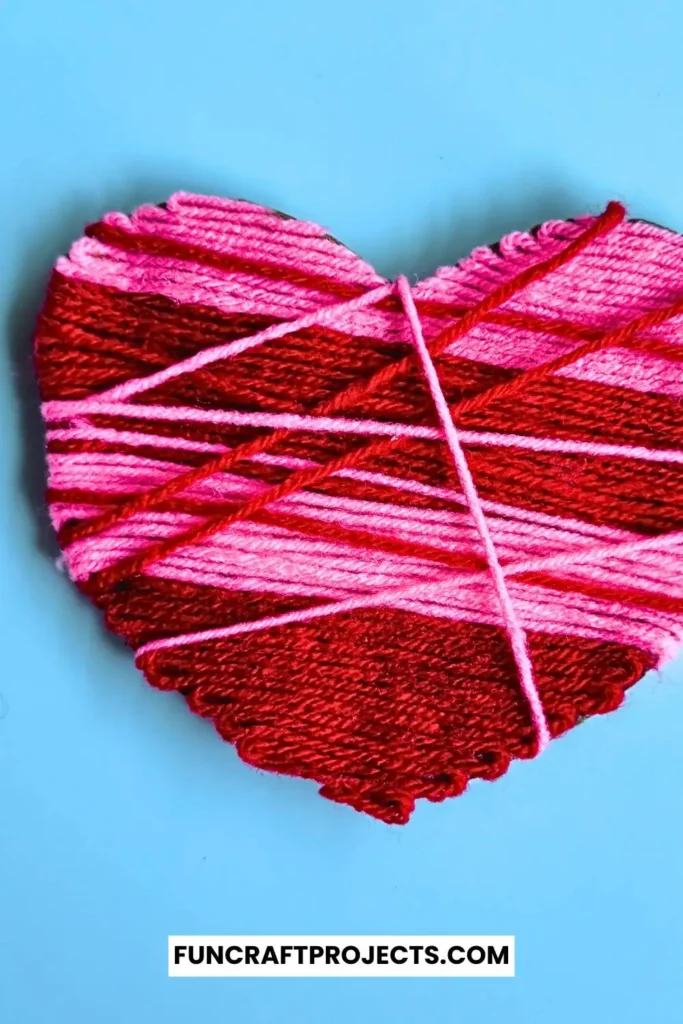 A vibrant yarn wrapped heart craft featuring pink and red yarn, wrapped intricately around a sturdy heart-shaped cardboard, perfect for Valentine’s Day.