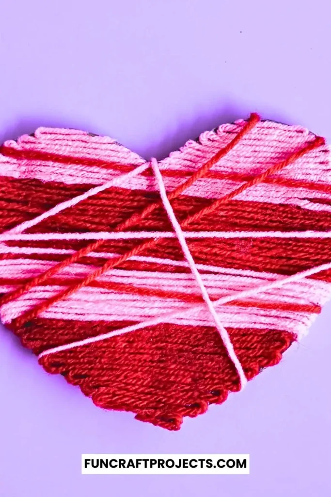 A vibrant yarn wrapped heart craft featuring pink and red yarn, wrapped intricately around a sturdy heart-shaped cardboard, perfect for Valentine’s Day.